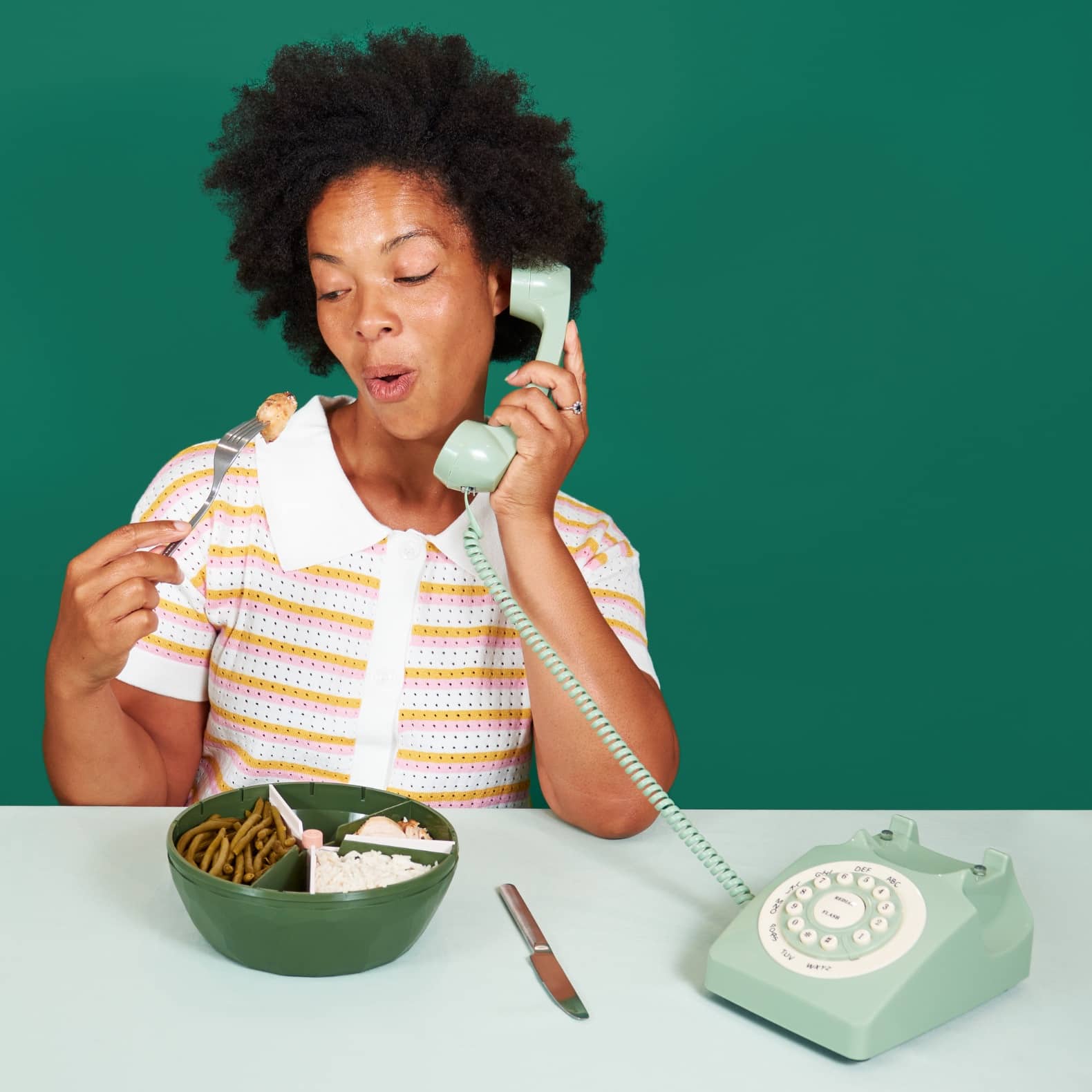 Fille qui téléphone avec un téléphone rétro de couleur verte, tout en mangeant avec une fourchette dans un bol maju de couleur kaki.